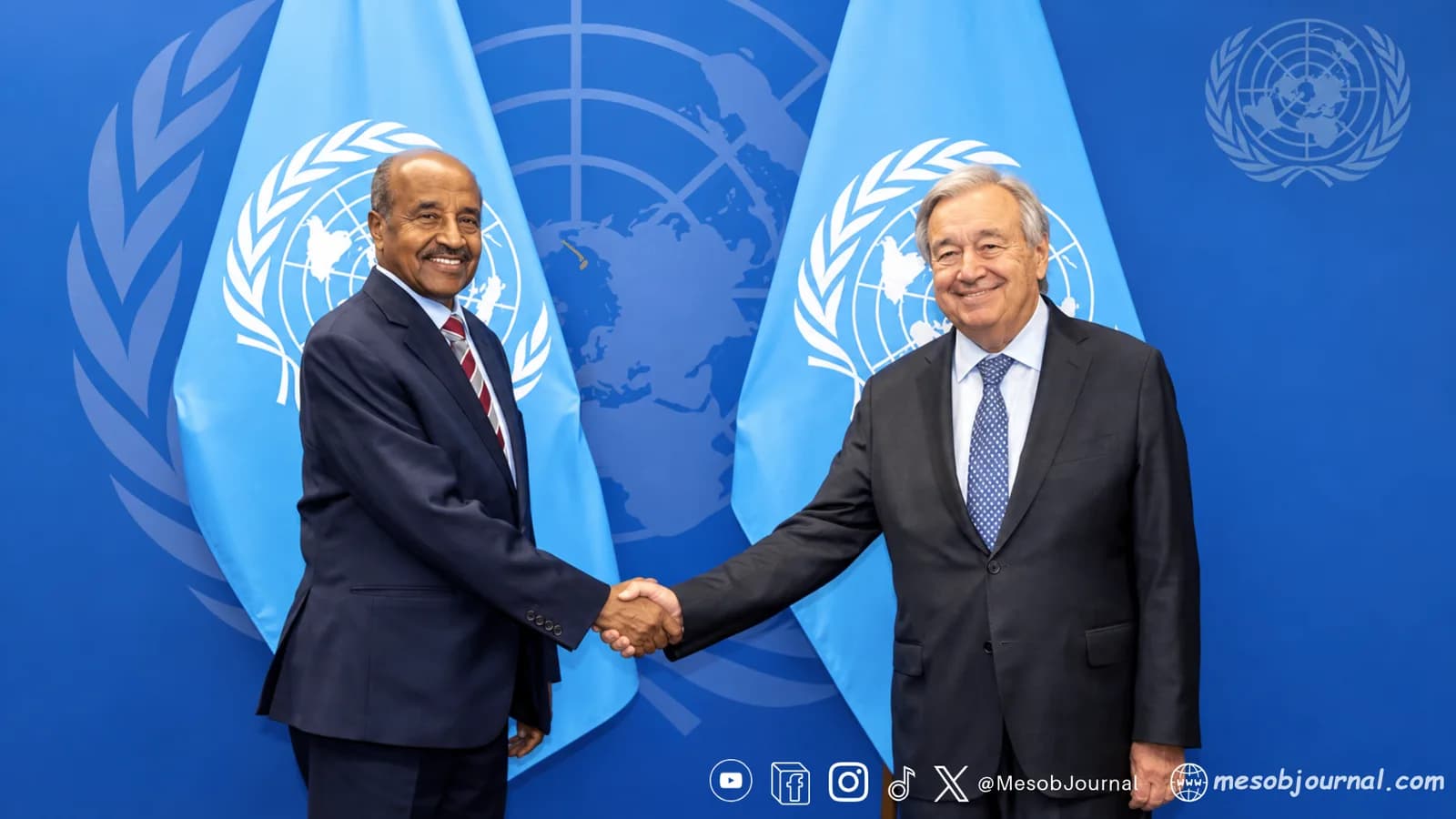 Two officials shake hands in front of United Nations flags.